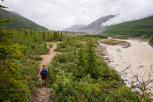 Researcher walking down a trail next to a river in a valley surrounded by mountains and fog. 