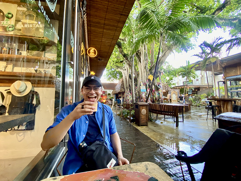 David sitting at a cafe table outside holding a cup of coffee and smiling. 