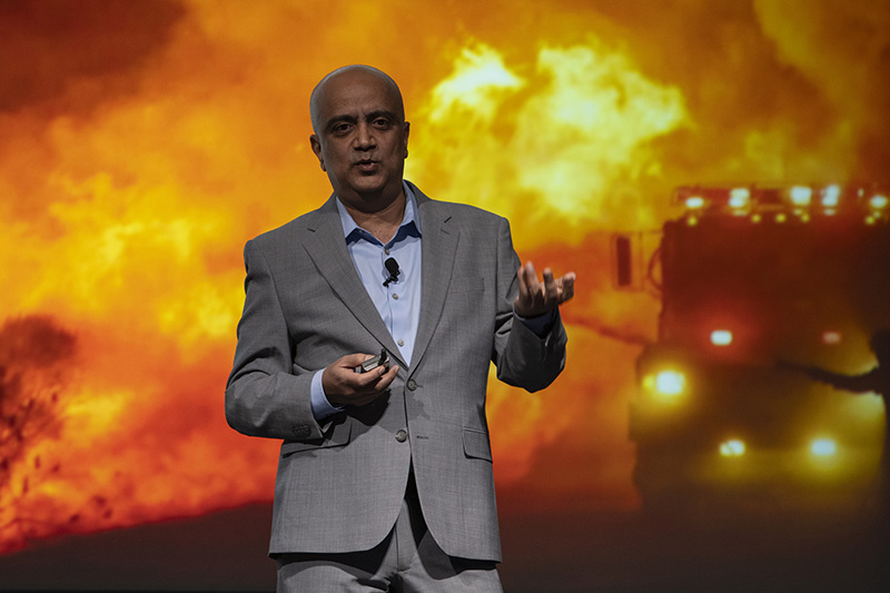 Dr. Kumud Acharya speaking on stage in front of a screen displaying a wildfire scene. 