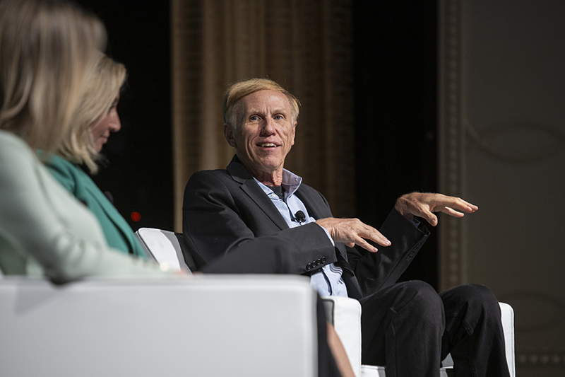 Dr. Tim Brown sitting in a white chair on stage with other panelists at the AWE+ Summit