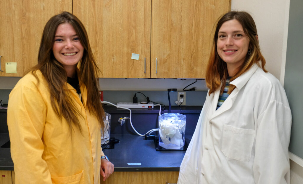 Photo of DePauw and Hines in lab coats standing in front of their experiment. 