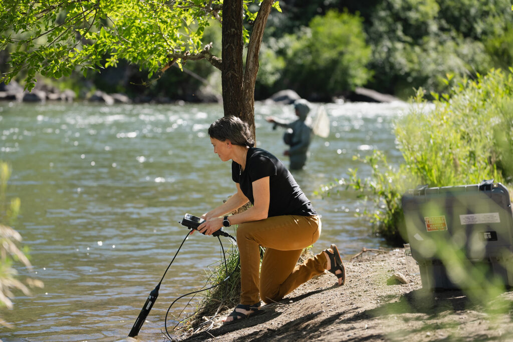 Researcher kneeling down at the edge of a river collecting water samples.