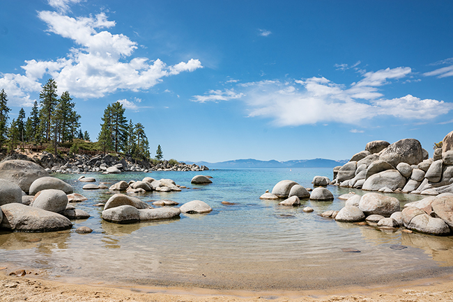 Crystal clear water on the coast of Lake Tahoe with boulders lining the beach and pine trees in the distance. 